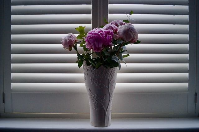 a vase of flowers stands on the windowsill with closed wooden blinds